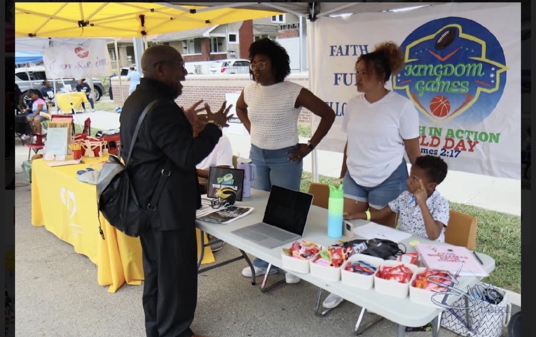 Huddle volunteers talking with a visitor at the Kingdom Games information booth beside the Faith in Action Field Day banner