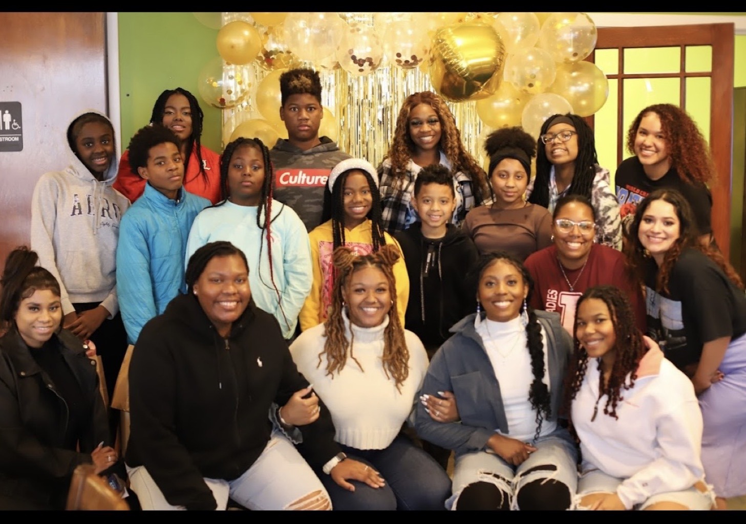 A large group of teens and young adult mentors gathered together at an indoor Huddle celebration with gold balloons