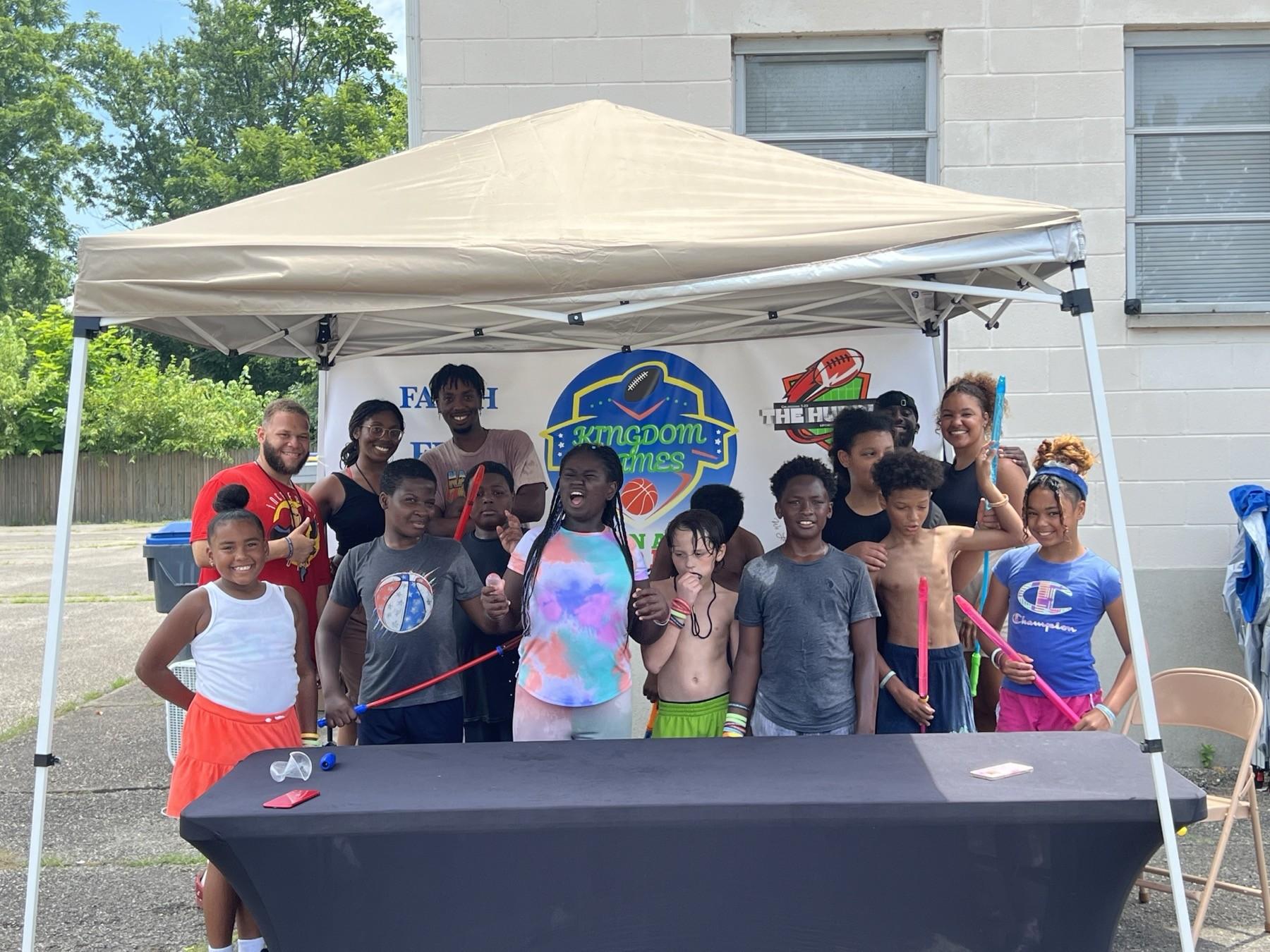 Youth and volunteers standing under the Kingdom Games tent with The Huddle banner at a community field day