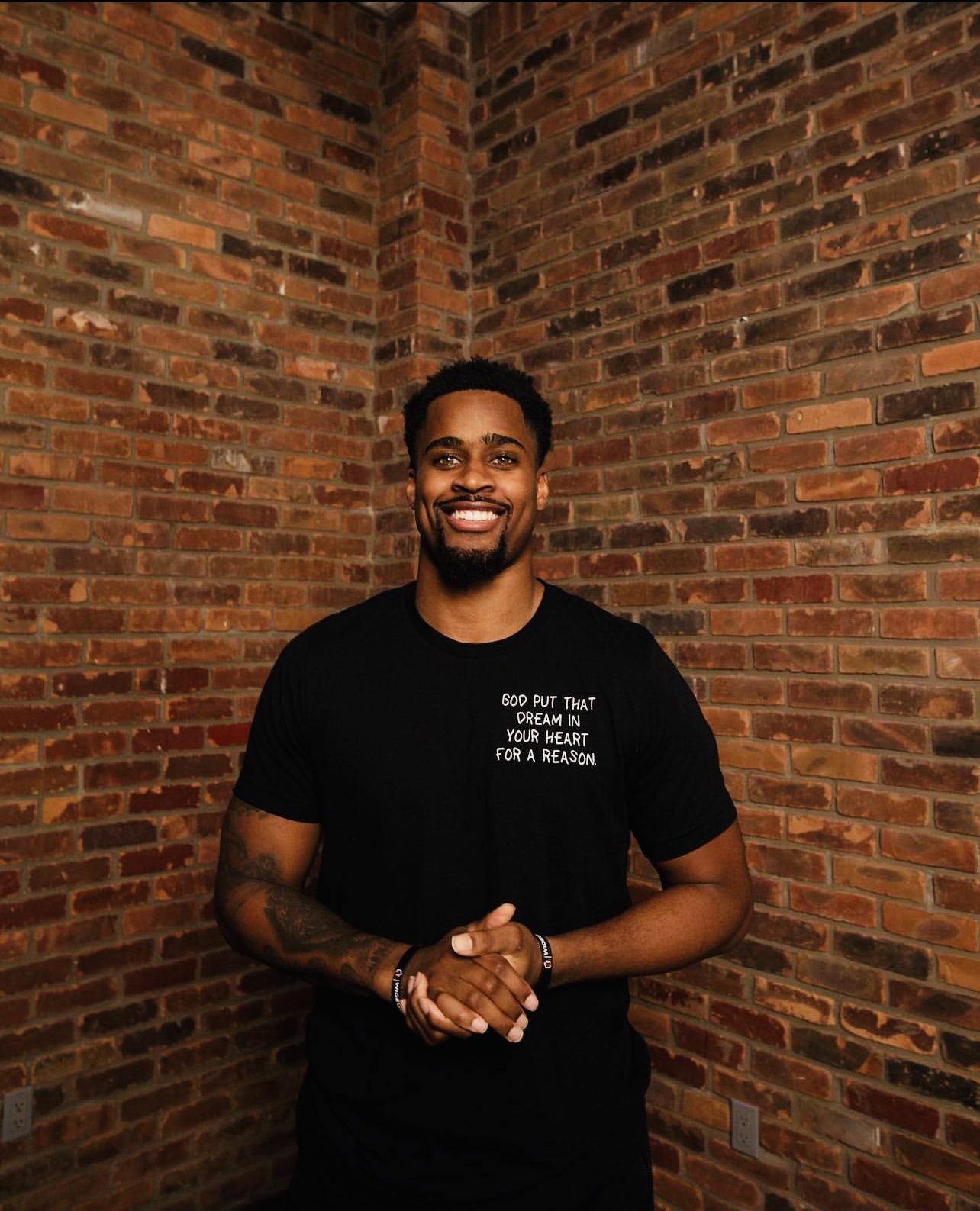Daylen Baldwin, CEO of The Huddle Inc., smiling in front of a brick wall wearing a black t-shirt