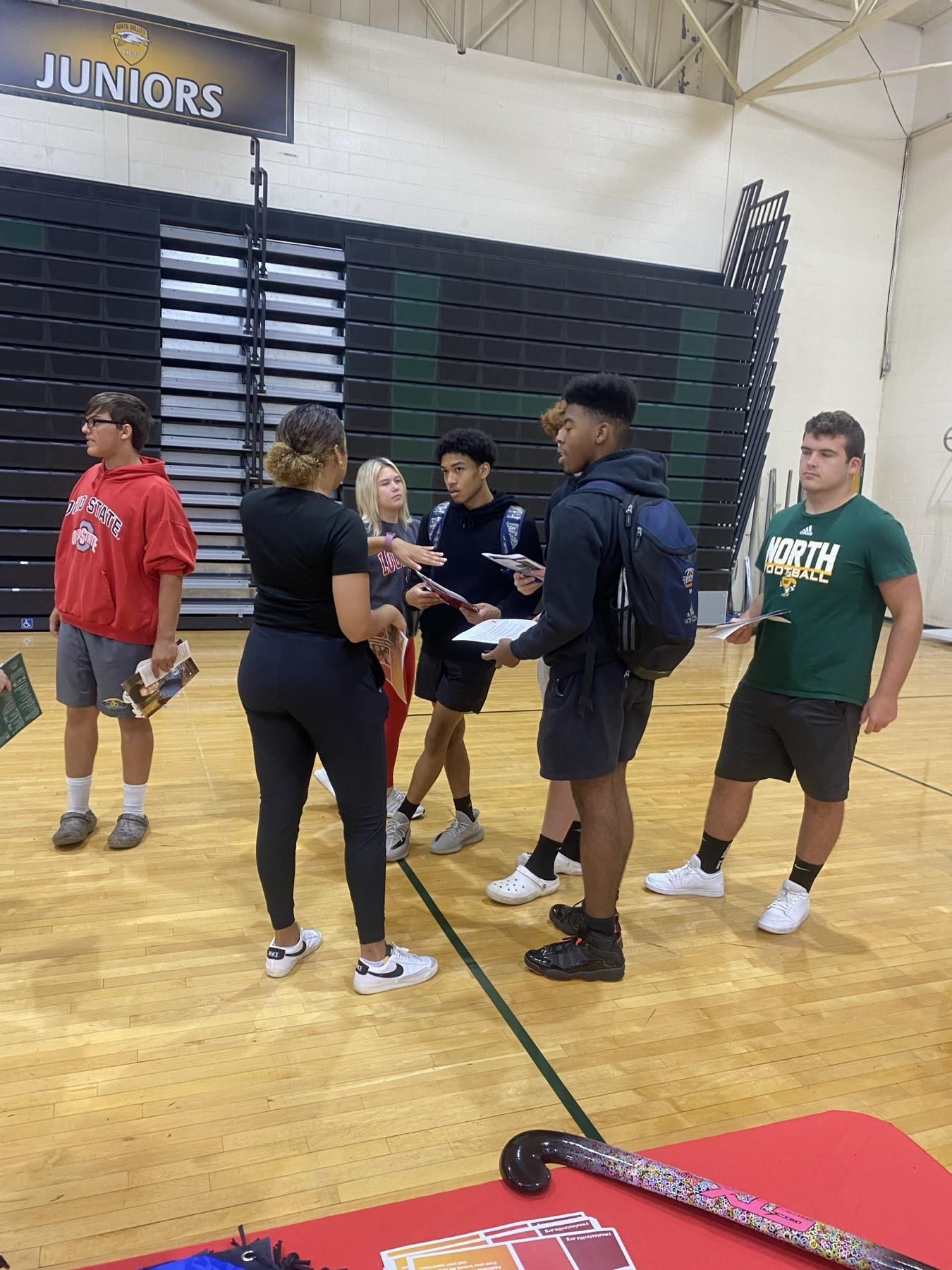 A Huddle leader talking with high school students at an outreach table in a school gymnasium