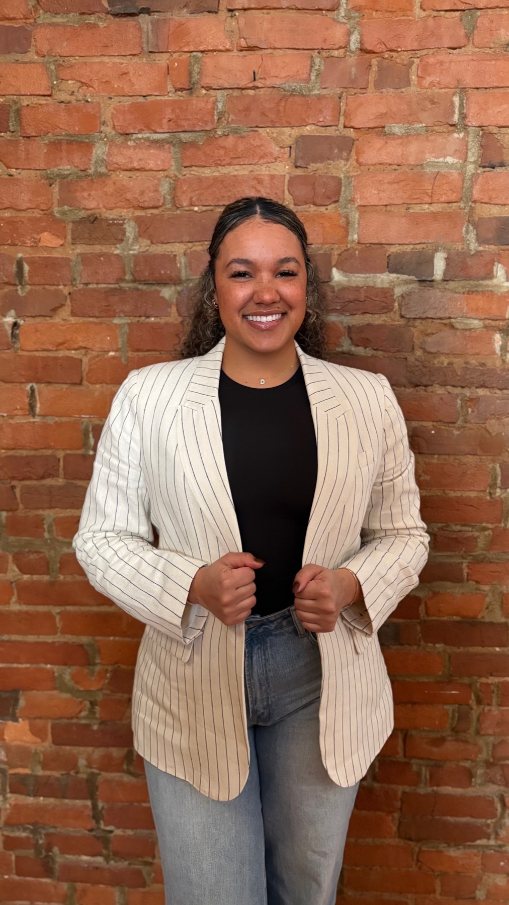 Kennadee Pringle, President and Founder of The Huddle Inc., smiling in a white striped blazer in front of a brick wall