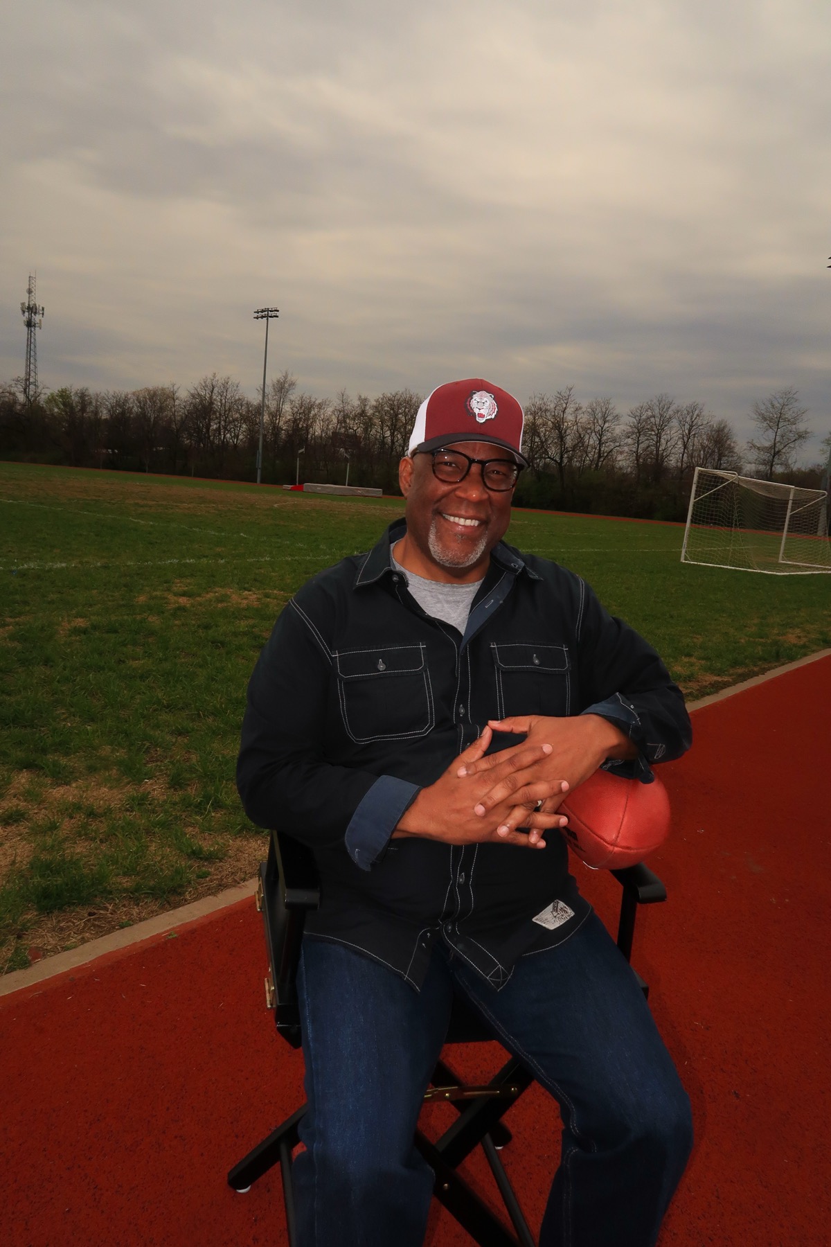 Kevin Pringle, Executive Director of The Huddle Inc., smiling while seated on a track holding a football